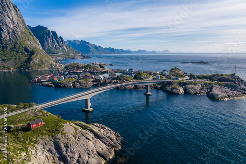 Obraz na płótnie Aerial view shows arching bridge linking rocky islets to Reine and Hamnoy in Lofoten, Norway, red rorbuer cabins, emerald slopes, gray rock, cobalt fjord and sea