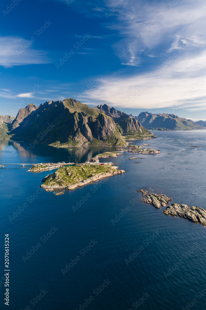 Fototapeta premium Aerial view of Lofoten Islands in northern Norway shows granite peaks above deep fjords, rocky islets, Reine and Reinebringen area, a coastal settlement, cabins, road, summer daylight