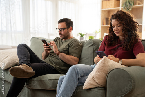 Obraz na plátně Angry young couple ignoring each other using phone after an argument while sitting on sofa at home