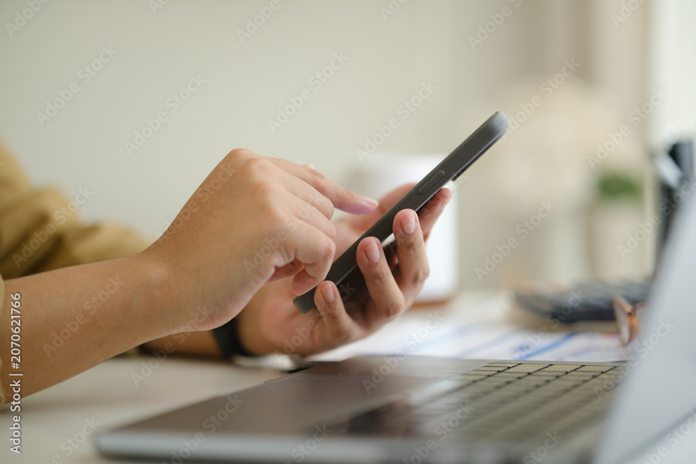 Fototapeta premium Close up of hands using smartphone beside laptop, calculator, and financial documents in a home office.