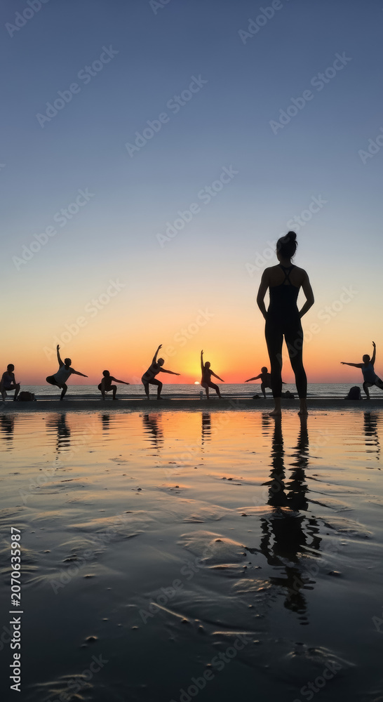 Fototapeta premium Yoga group practices mindfulness and physical exercise during a golden hour sunset on the reflective sandy beach shore.