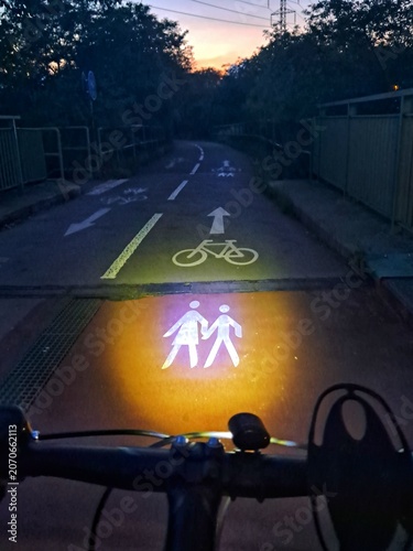 First person view of a bicycle handlebar at night with a bright headlight illuminating a pedestrian symbol on a dark path.