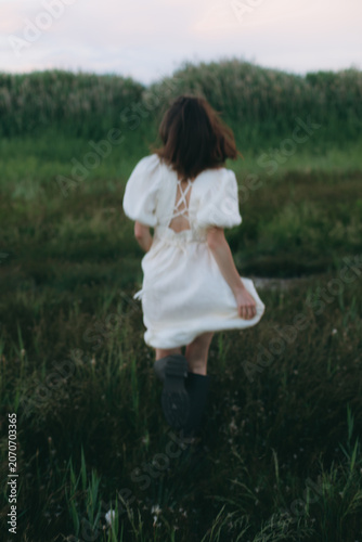 Dynamic brunette woman from back view running away genuinely into a green meadow field, wearing an authentic local white linen dress, freedom spirit atmospheric escape with creative motion blur
