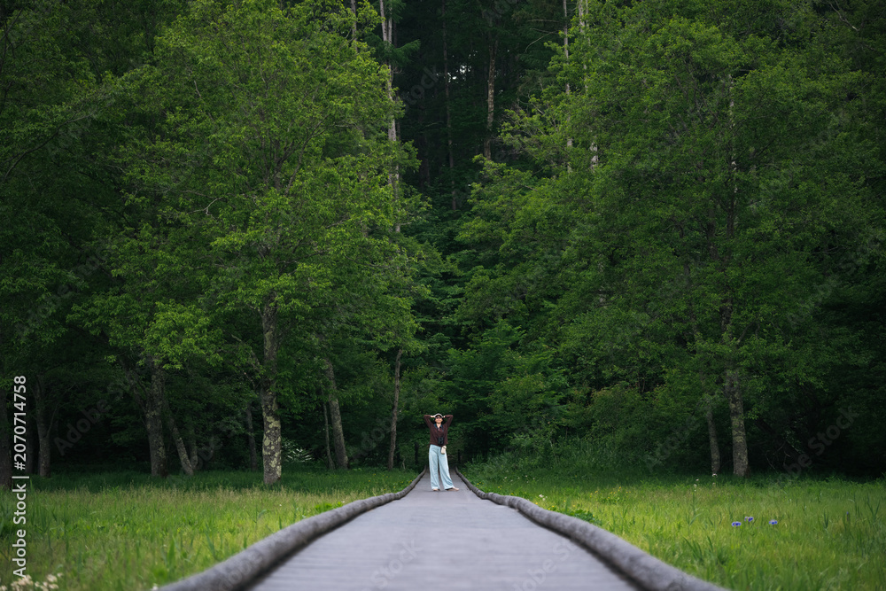 Fototapeta premium Forest boardwalk woman meadow tree green nature summer, lone woman standing on wooden path through lush woodland and grassy meadow with calm expression