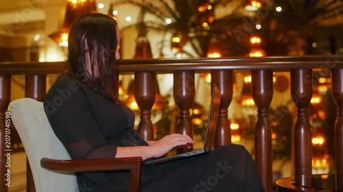 Woman sits at elegant balcony railing, typing on laptop amid warm golden bokeh lights, creating luxurious evening work atmosphere in sophisticated interior setting.