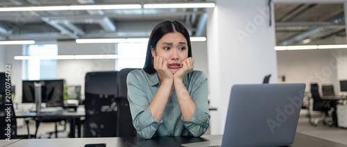 Stressed young Asian businesswoman looking at laptop screen in modern office, workplace anxiety, overwhelmed employee, corporate burnout.