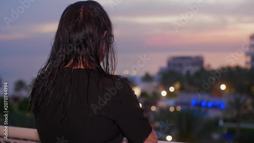 Woman with long dark hair viewing urban landscape at sunset from elevated vantage point, bokeh lights illuminating evening cityscape below.