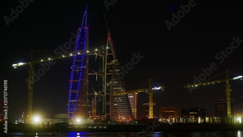 Night scene of industrial harbor development featuring illuminated construction cranes, modern office buildings, blue architectural lighting, reflected in dark water.