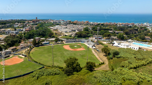 Photos Aerial panorama of a beautiful baseball field nestled within a coastal sports complex, showcasing the green outfield, red dirt infield, and a stunning blue sea horizon