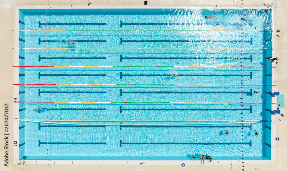 Obraz premium Aerial top down view of a bright blue swimming pool divided into multiple lanes having colorful floating markers, showing several athletes training together during a sunny summer day. Zenithal shot.