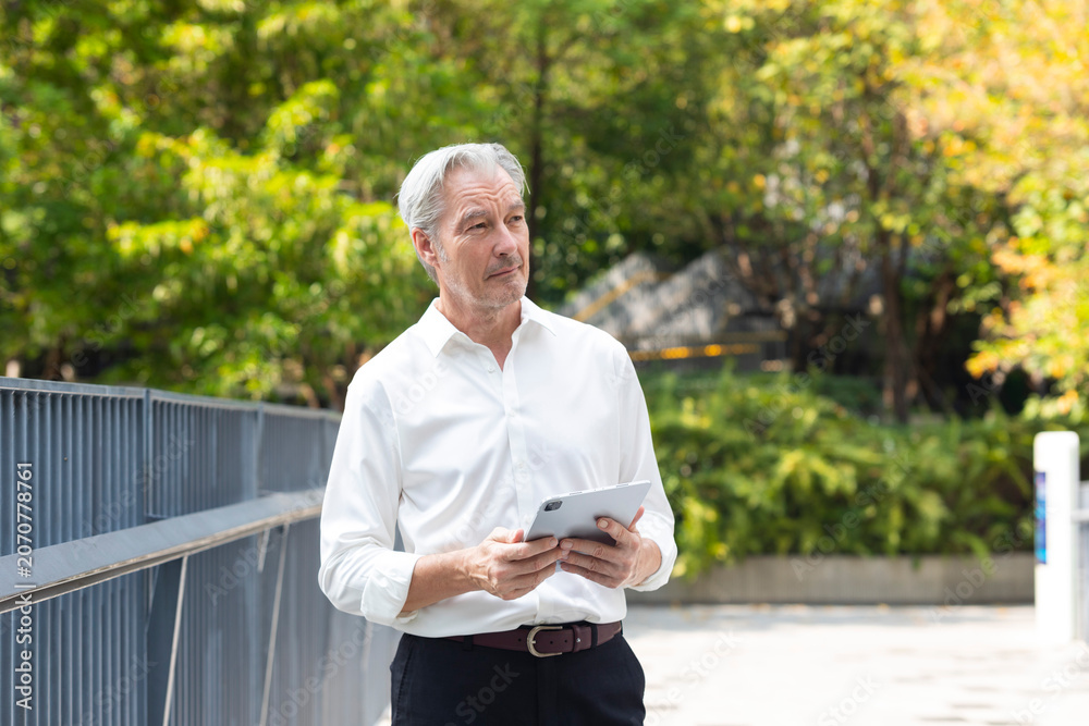 Obraz premium Smart and handsome senior businessman in white shirt checking email with tablet in outdoor office space and park. Business and digital technology concept.