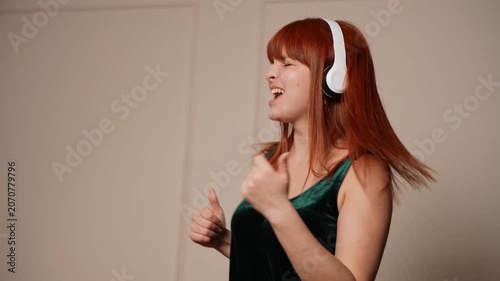 Happy emotional young woman listening to music in wireless headphones at home. Cheerful redhead girl singing and dancing in living room. Domestic leisure, stress relief and active youth lifestyle