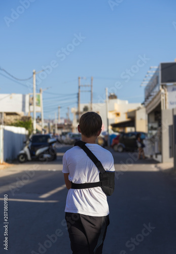 Rear view of an 11-year-old boy with a broken arm in a black medical sling walking down a street during a summer vacation in Kato Gouves, Crete, Greece