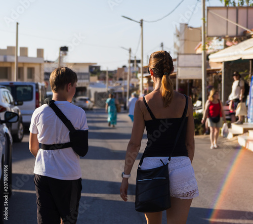 Rear view of a mother walking down a sunny street with her 11-year-old son who has a broken arm in a medical sling during a summer vacation in Kato Gouves, Crete
