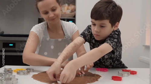 Mother and son cut shapes from gingerbread dough with metal cutters at kitchen table. Family prepares sweet homemade cookies for holiday baking recipe education lifestyle leisure context.