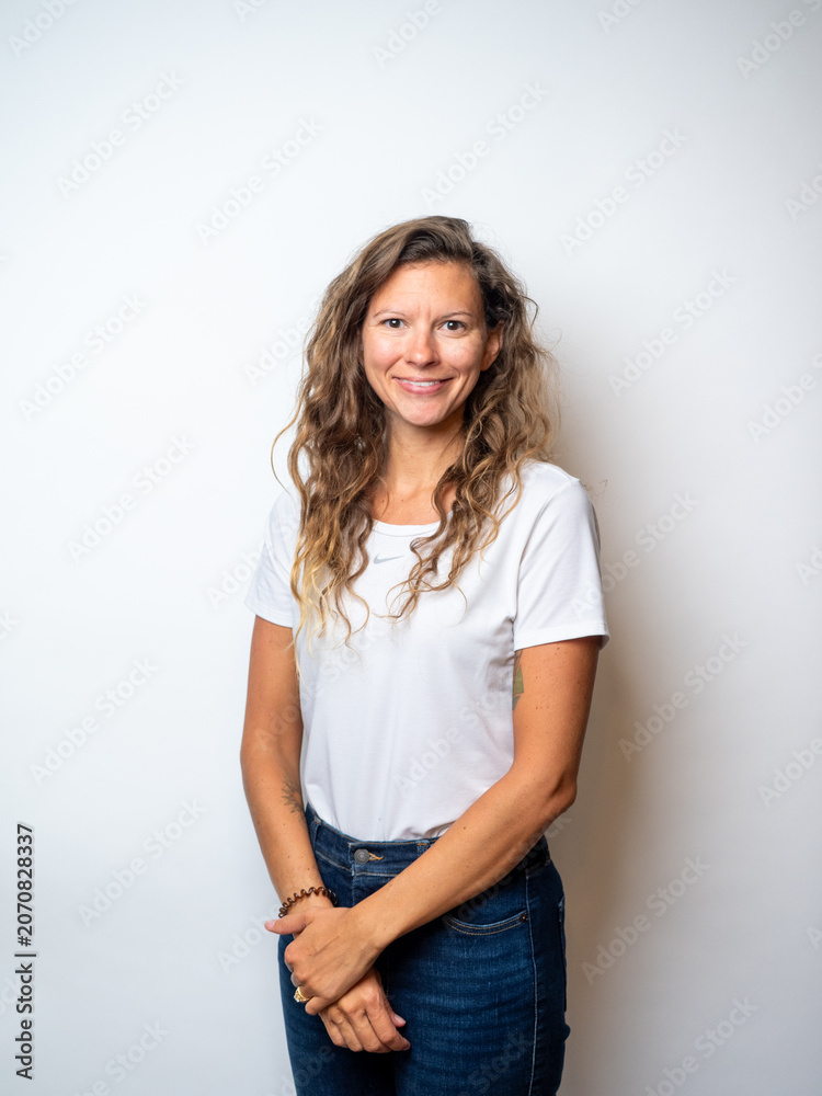 Fototapeta premium Studio portrait of a mid-thirties woman on a white backdrop wearing a white t-shirt and dark blue jeans, with dark blonde wavy hair and a happy, smiling expression.