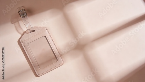 Pale pink luggage tag with a clear window and transparent buckle strap rests gently on a soft, light-colored, quilted surface indoors. The light casts subtle shadows