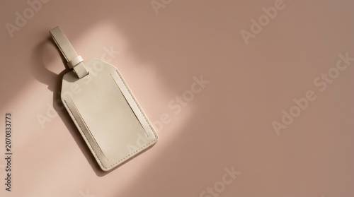 A clean, light beige luggage tag lies on a muted pink background, illuminated by soft natural light creating a gentle shadow. This accessory suggests travel and organization for a seamless journey