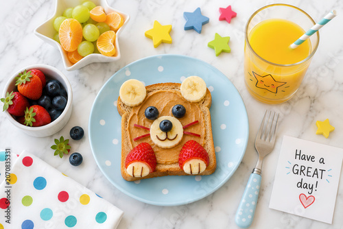 A kitchen table displays a fun breakfast with toast shaped like a bear, topped with fruit. There is a glass of juice and a bowl of fresh berries and other fruits nearby.