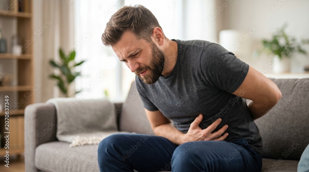Fototapeta premium A man sits on a couch holding his stomach with an expression of discomfort. He looks concerned while sitting in a well-lit living room with plants and a cozy atmosphere
