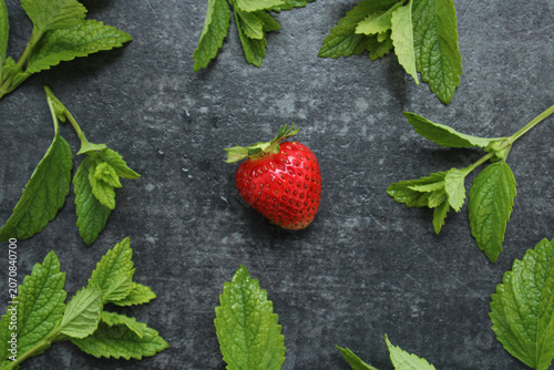 Single ripe red strawberry centered on a dark textured slate background, framed by fresh green mint leaves in a minimalist top-down flat lay composition.