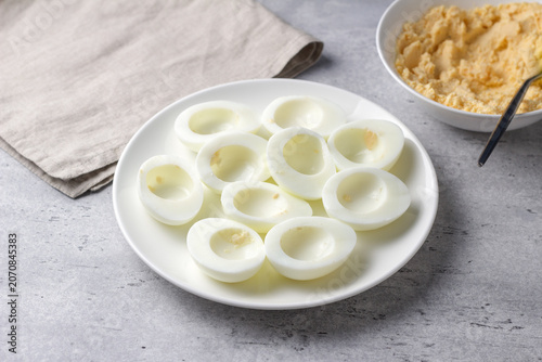Hard boiled egg halves with yolks removed on a white plate, prepared for stuffed eggs recipe on a gray textured surface with soft natural light