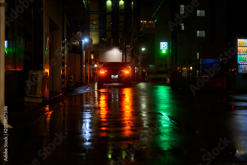A moody nighttime photograph of a narrow wet street in Japan, illuminated by colorful artificial lights and their reflections on rain-soaked pavement. In the center, a dark-colored car with glowing