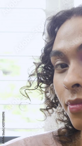 Vertical video: Looking cam nonbinary dabbing paint on cheek near blinds checking brush, copy space