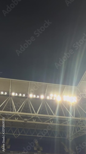 Stadium floodlights brightly illuminating the night sky over a modern sports arena, highlighting the powerful architectural structure of the grandstand roof and beams Football stadium with floodlights