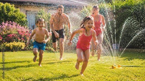 Family of parents and two children running happily through a backyard lawn sprinkler on a sunny summer day, playing and splashing in water