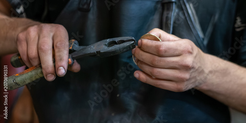 Fotografie A craftsman wearing a black leather apron uses a pair of old hand pliers to carefully hold and manipulate a small round brass or metal object in a workshop
