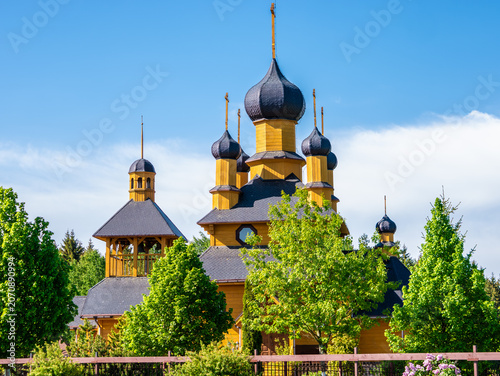 Foto A traditional yellow wooden Orthodox church featuring multiple dark onion domes and a bell tower stands behind green summer trees against a blue sky