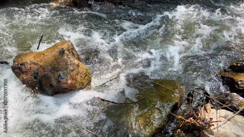Fast flowing river stream rushing around mossy rocks with white foam and fallen leaves on bank