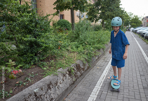 Young boy in helmet rides a skateboard on a paved path next to green bushes