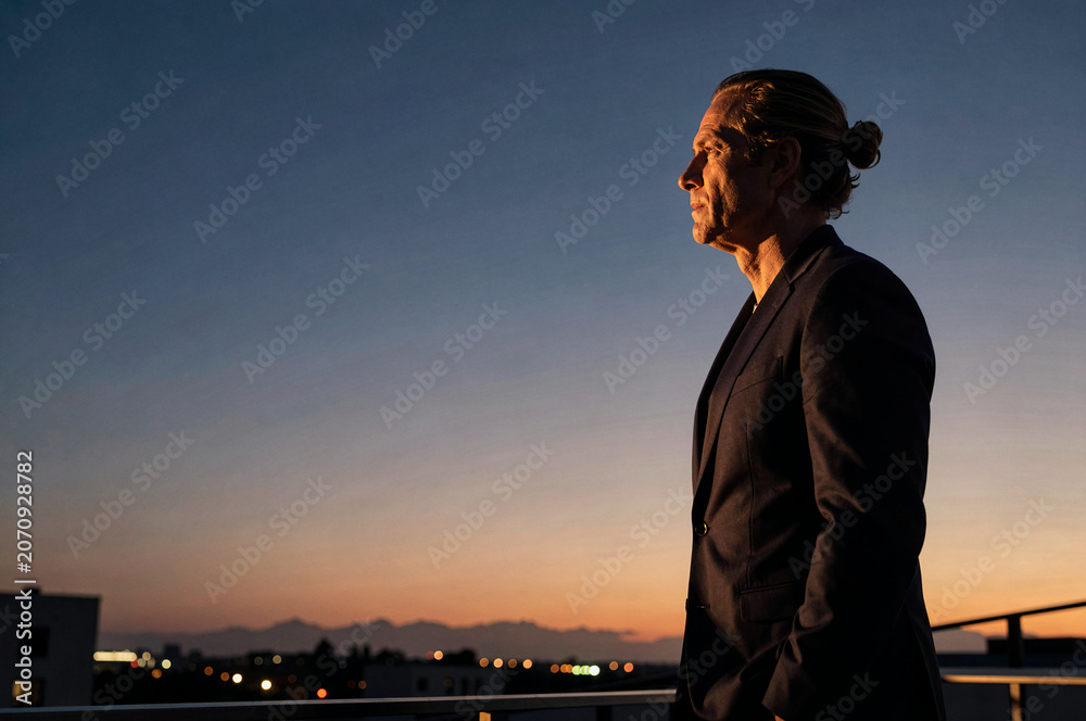 Fototapeta premium Man in dark suit stands on a balcony during twilight. Golden hour light illuminates his profile against an orange and blue gradient sky over distant city lights and mountains.