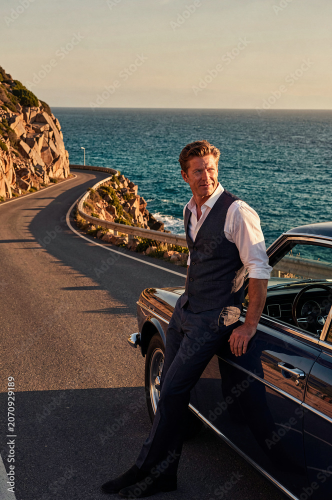 Fototapeta premium Man in formal waistcoat leans against a classic dark blue convertible car along a coastal road. Warm sunset light hits his face while the ocean waves crash nearby rocky cliffs.