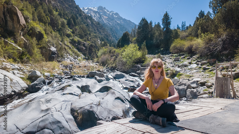 Fototapeta premium A young woman sits peacefully by a flowing river, surrounded by majestic mountains and lush greenery.