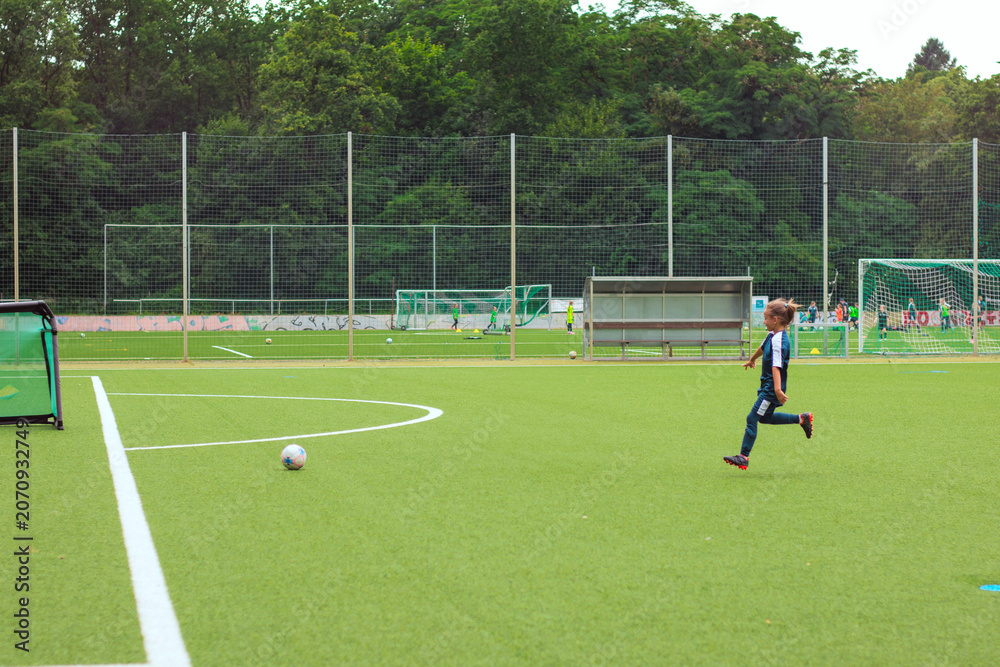 Obraz premium Young boy in sportswear runs towards a soccer ball on a green artificial turf field with goalposts and players visible in the background