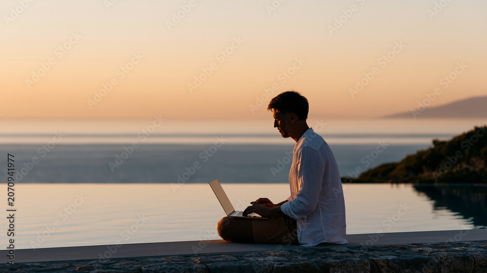 Fototapeta premium Young man sitting poolside working on a laptop at sunset, tropical vacation freelance remote online work concept, with copy space