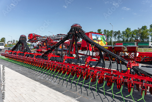 Fotografie A long red soil cultivator with multiple metal tines and black hydraulic hoses parked on gravel in a farm equipment lot
