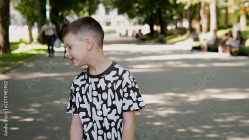 Boy in patterned black and white t-shirt stands on sun-dappled paved park pathway, head turned sideways with slight smile, surrounded by mature trees, benches