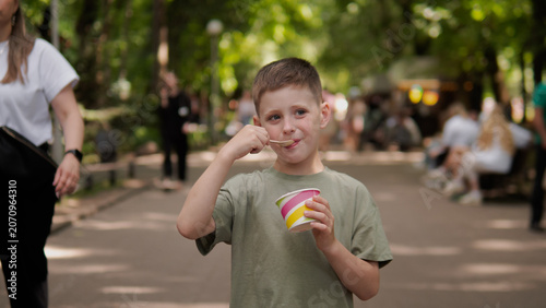 Smiling young boy in casual t-shirt enjoys cup of colorful ice cream while standing on paved path of shaded urban park; soft sunlight filters through leafy