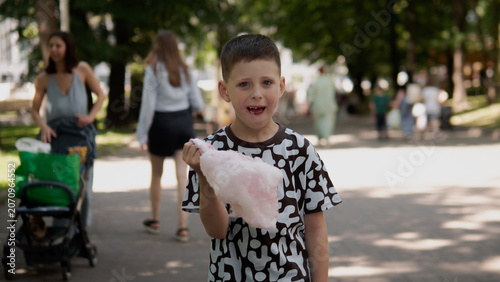 Young boy stands on tree-lined park pathway holding pink cotton candy while smiling toward camera; casual summer clothing, patterned T-shirt, short haircut