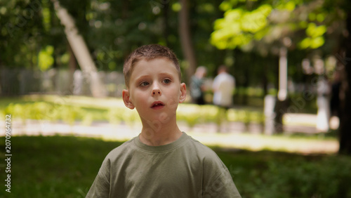 Close-up portrait of young boy standing in sunlit city park, wearing olive green shirt, with short brown hair, wide eyes, open mouth, and surprised face