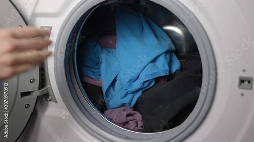 Woman's hand loading dirty clothes into a washing machine. Close-up view of a person's hand putting colorful laundry into the drum of a modern front-loading washing machine, then closing the door