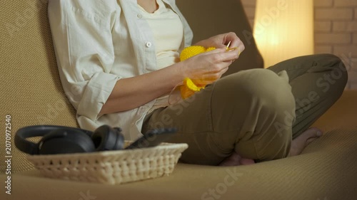 Woman knitting while relaxing on the couch. Relaxed woman sitting on a comfortable sofa in the living room, knitting with yellow wool yarn to create a handmade garment, enjoying her quiet hobby