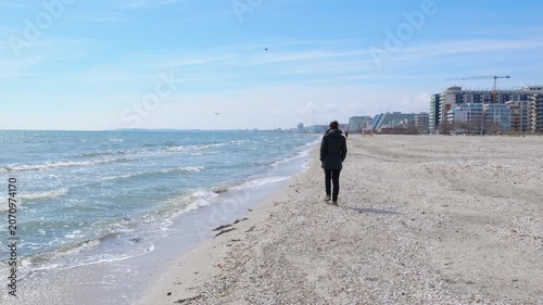 Solitary person walking along a sandy beach. Melancholic person walking alone along the seashore on a cold, sunny day with the city skyline in the background, enjoying a moment of solitude