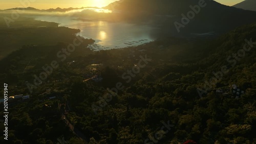 Aerial view of Lake Batur and Mount Abang at sunrise with golden light reflecting on the water and mist over the lush volcanic slopes in Kintamani, Bali, Indonesia.