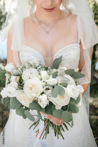 Lovely bride wearing an elegant white wedding gown and veil while holding a lush bouquet of white roses, delicate seasonal flowers and eucalyptus foliage, standing outdoors during wedding celebration
