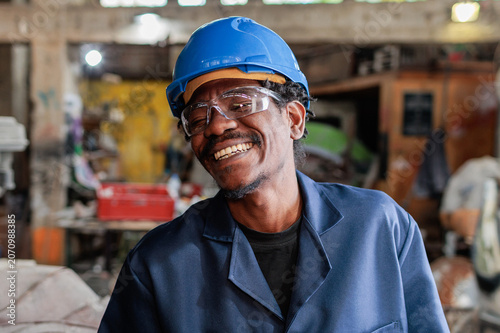 African worker at construction site wearing a blue hard hat and safety goggles, smiles while surrounded by tools and equipment